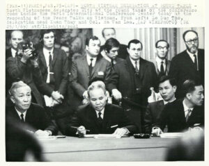 A black and white photo shows officials sitting and standing around a table as one man in the middle signs a piece of paper